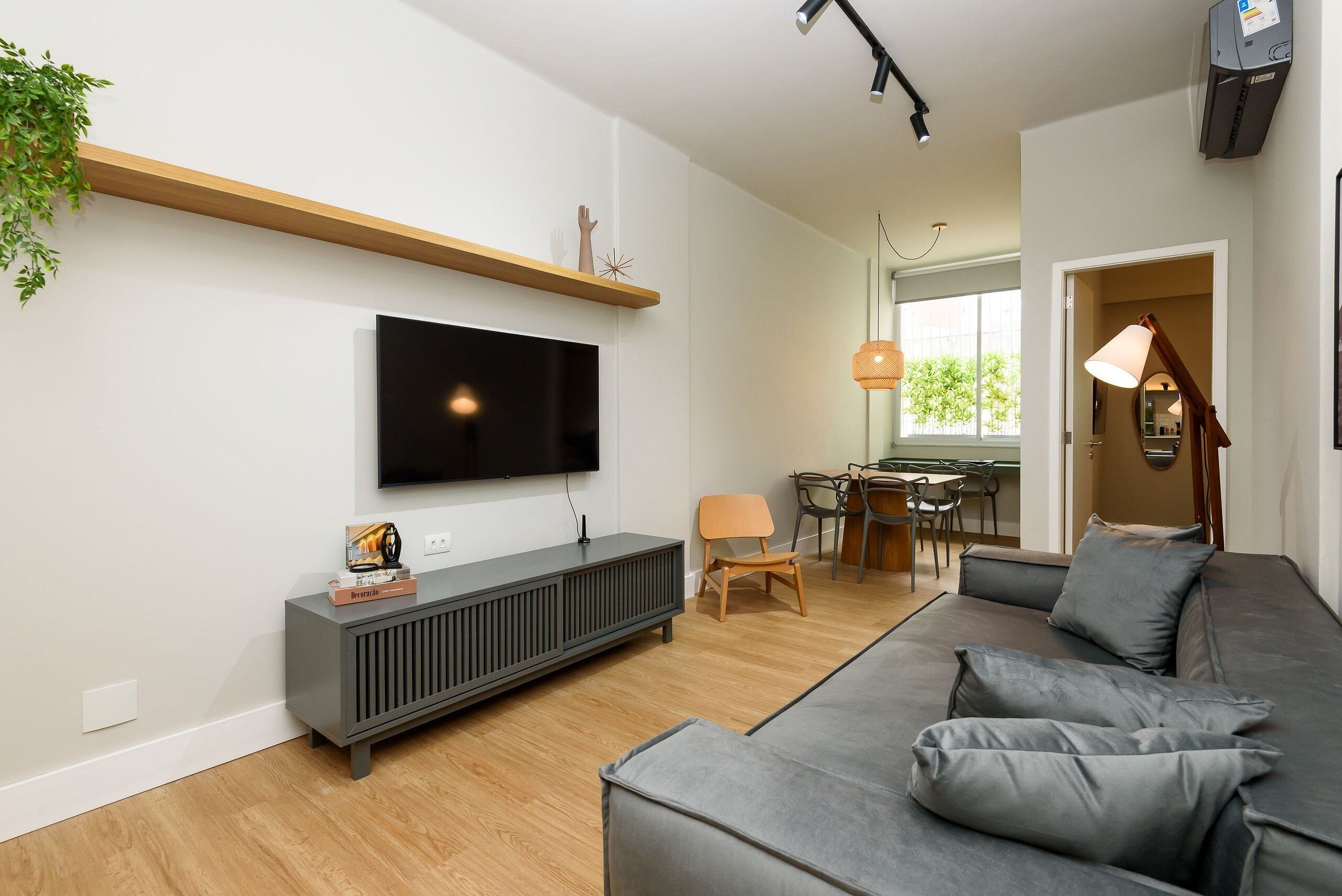 A modern living room with a grey sofa, a flat-screen TV, a wooden shelf, and a dining area with chairs and a table.
