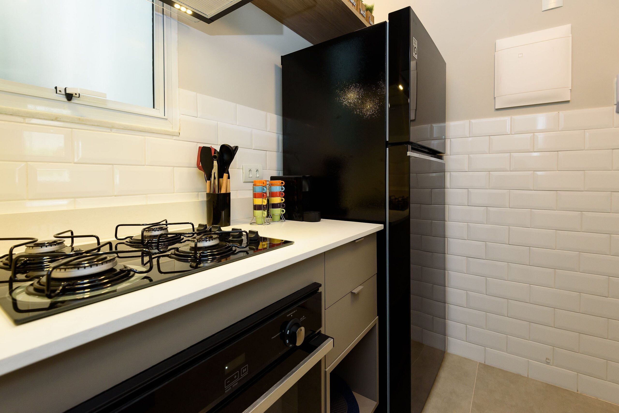 Una cocina con estufa a gas, refrigerador negro, salpicadero de azulejos blancos y una ventana sobre la estufa.