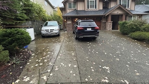A residential driveway with two cars parked, a stone wall, and a house with a gabled roof.