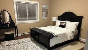 A bedroom with a bed, a desk with a mirror, a window with blinds, a lamp, and a rug.