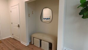 Un pasillo con una puerta blanca, un banco, un espejo y una planta.