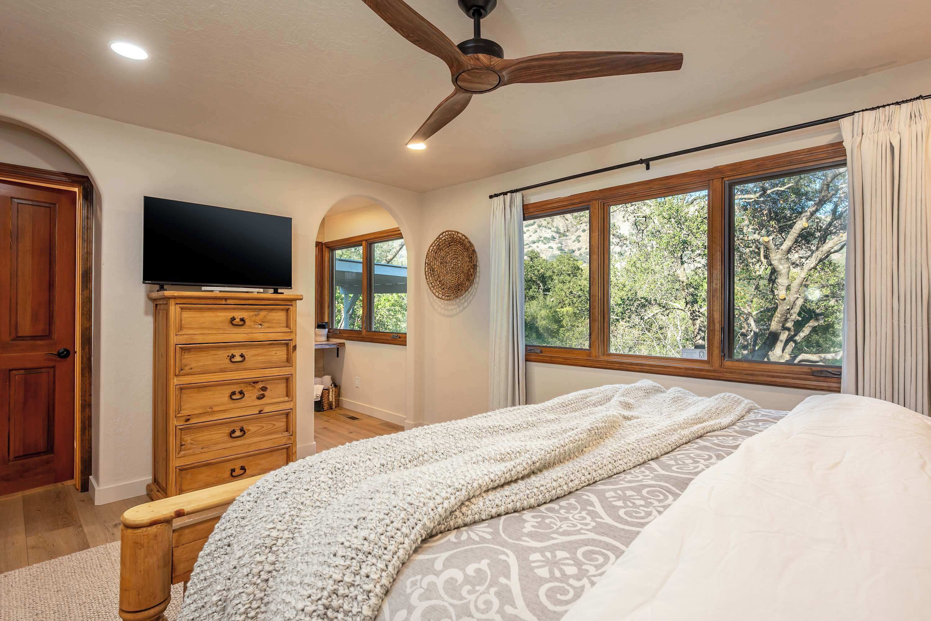 Une chambre à coucher comprenant un lit, une commode en bois, une télévision, un ventilateur de plafond et de grandes fenêtres donnant sur la forêt.