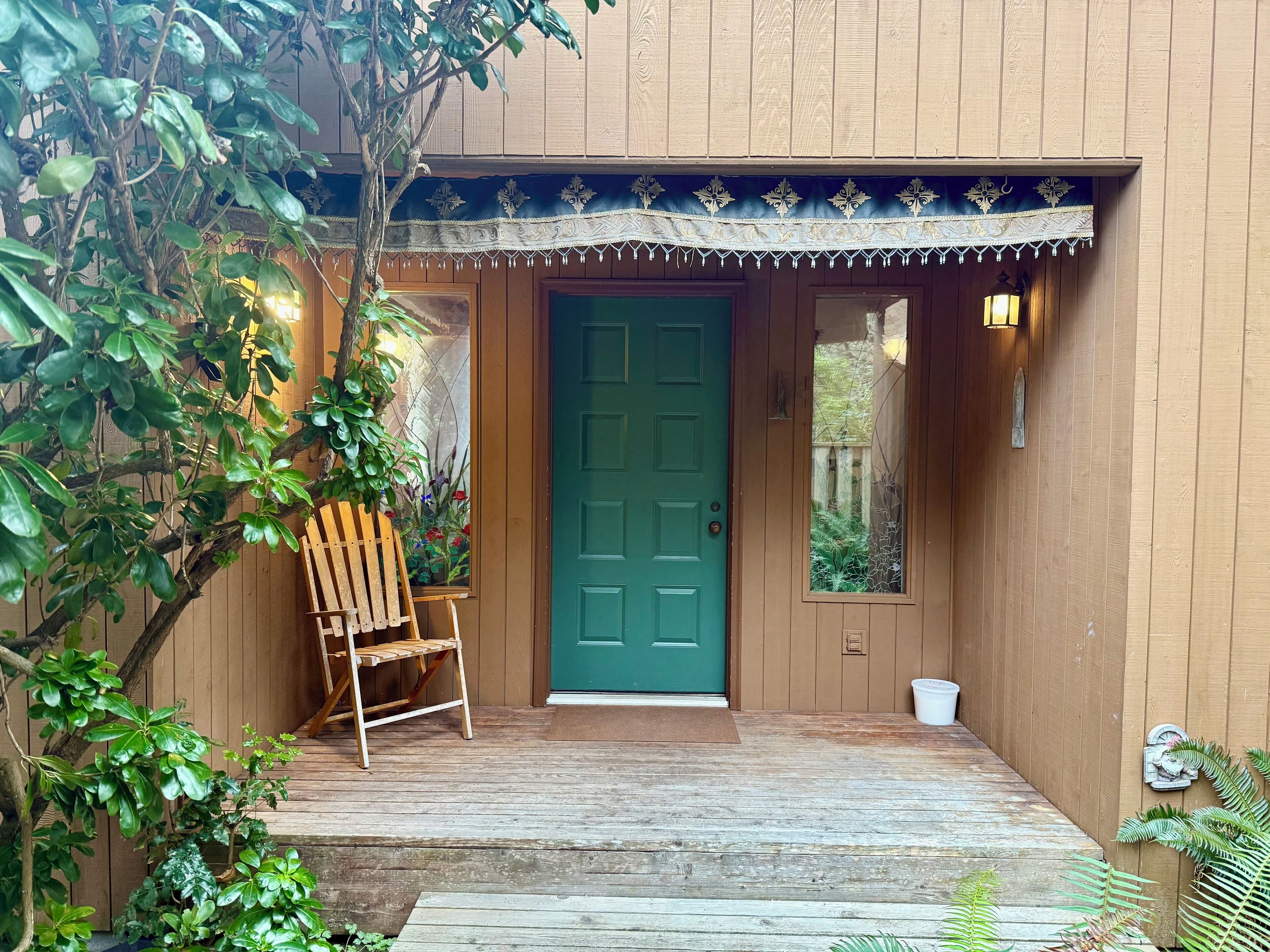 Una construcción de madera con una puerta verde, una silla de madera afuera y un pequeño jardín.