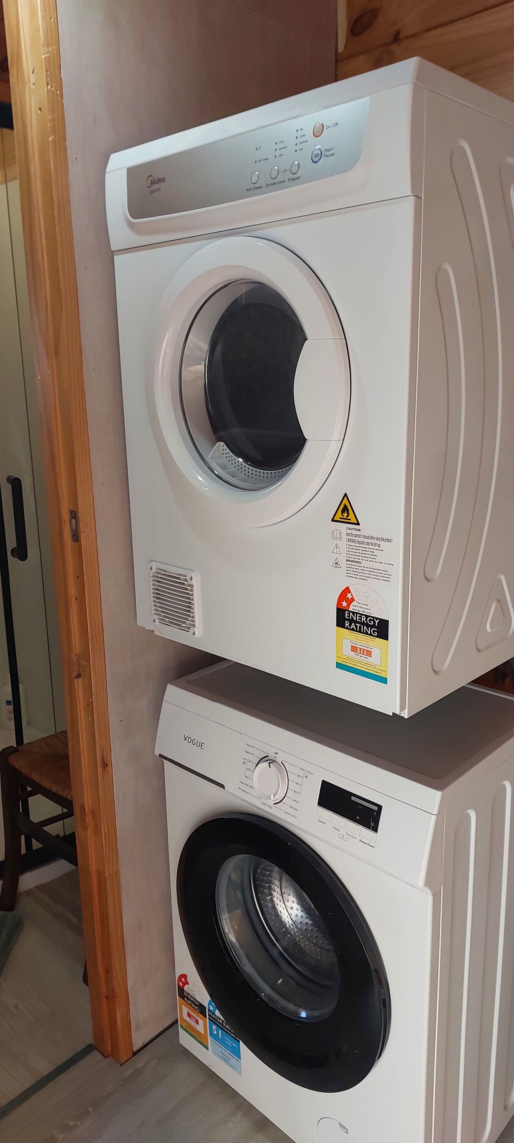 Two stacked front-loading washing machines, one on top of the other, in a room with wooden flooring and a door visible in the background.