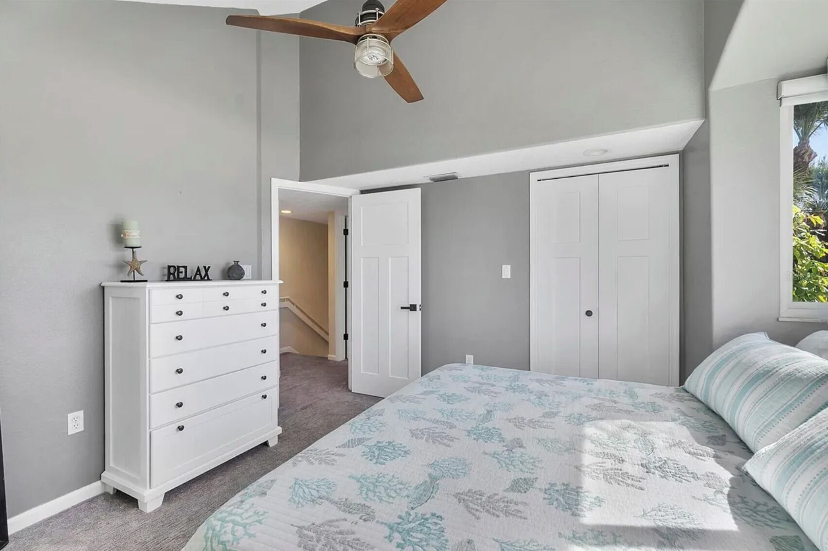 A bedroom with a bed, a dresser, a ceiling fan, and a window with a view of greenery.