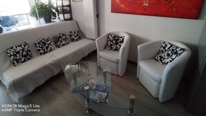 A modern living room with a white sofa, two white armchairs, and a glass coffee table. A red abstract painting adorns the wall, and a potted plant is visible near the window. - Apartment T2 45M2 exceptional view Fitted kitchen shower room living room loggia (Agde)