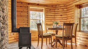 Interior de una cabaña de madera con pared de piedra, estufa de leña negra, mesa de comedor de madera, sillas y ventanas grandes.