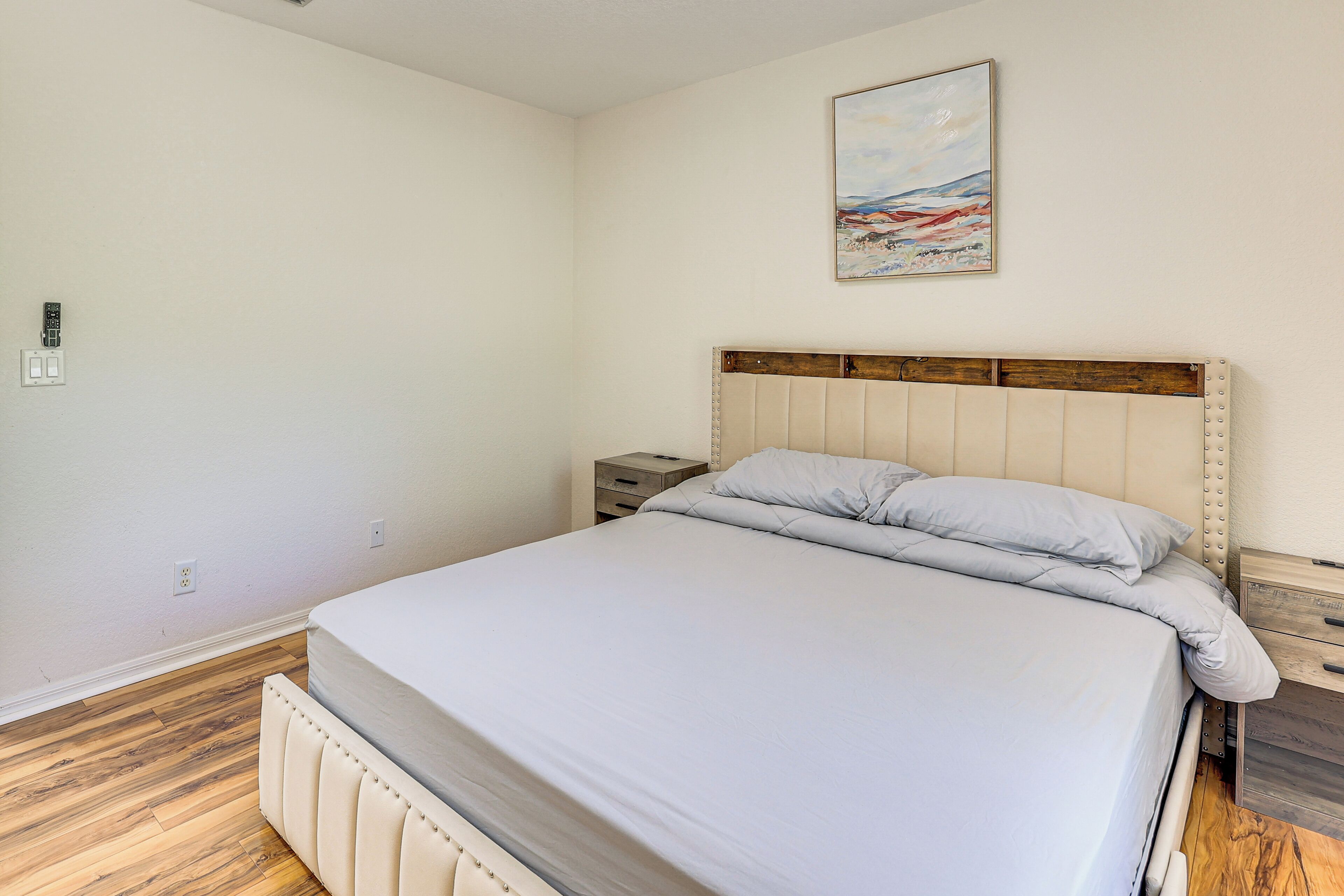 A bedroom with a bed, wooden flooring, a nightstand, and a painting on the wall.