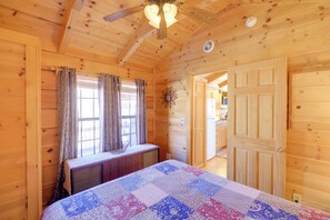 Une pièce de cabane en bois comprenant un lit, un ventilateur de plafond, une fenêtre avec des rideaux et une porte.