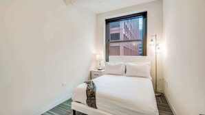 A hotel room with a large bed, a nightstand, a lamp, and a view of buildings through a window. - Downtown Chicago Stay with Free In & Out Parking 3 (Chicago)