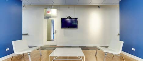 Un área de espera con dos sillas blancas, una mesa blanca, un televisor montado en la pared y una puerta de salida.