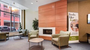 A lobby with patterned armchairs, a round table, a fireplace, and a large window with a view of a building and parked cars.