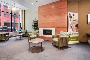 A lobby with patterned armchairs, a round table, a fireplace, and a large window with a view of a building and parked cars.