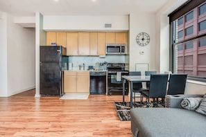 A compact kitchen with wooden cabinets, a black refrigerator, a microwave, and a stove. There is a dining area with a table and chairs, and a large window with blinds. - Downtown Chicago Stay with Free In & Out Parking 3 (Chicago)