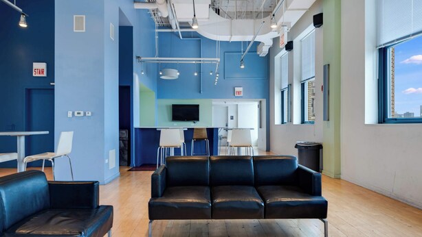 A modern lounge area with a black leather sofa, a small table, and chairs. There is a television mounted on the wall and an exit sign above the door.