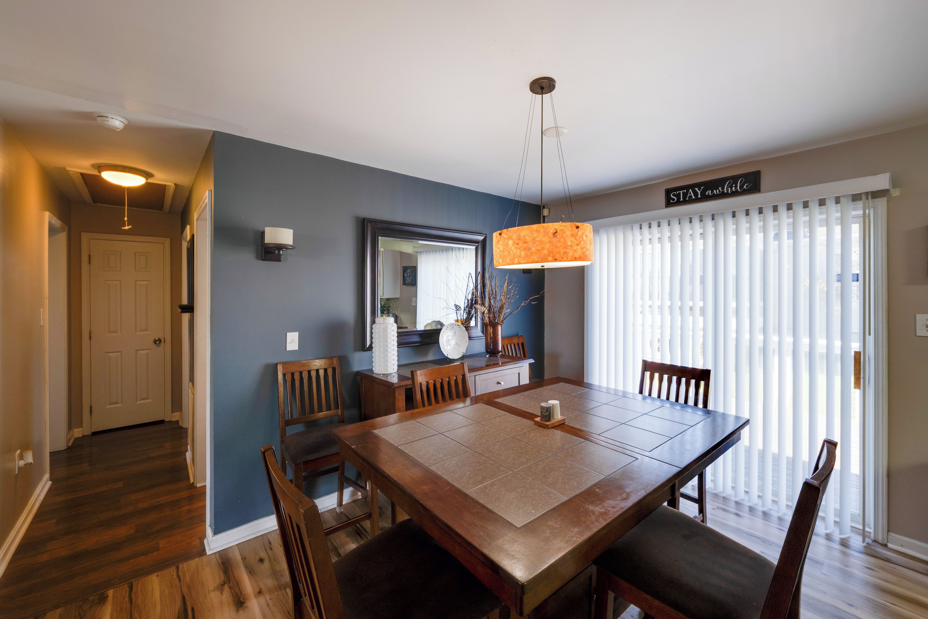 A dining area with a wooden table and chairs, a pendant light, a mirror, and a sign that reads "STAY awhile".