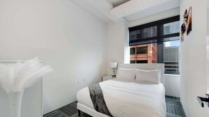 Una habitación de hotel moderna con una cama grande, una mesita de noche, una lámpara, un jarrón con plumas blancas y una ventana con vistas a un edificio.