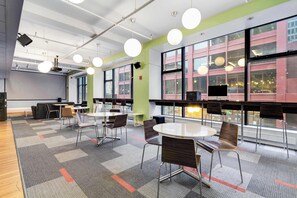 Une salle de conférence moderne, dotée de tables rondes, de chaises et de grandes fenêtres offrant une vue sur un bâtiment à la façade rouge et blanche.