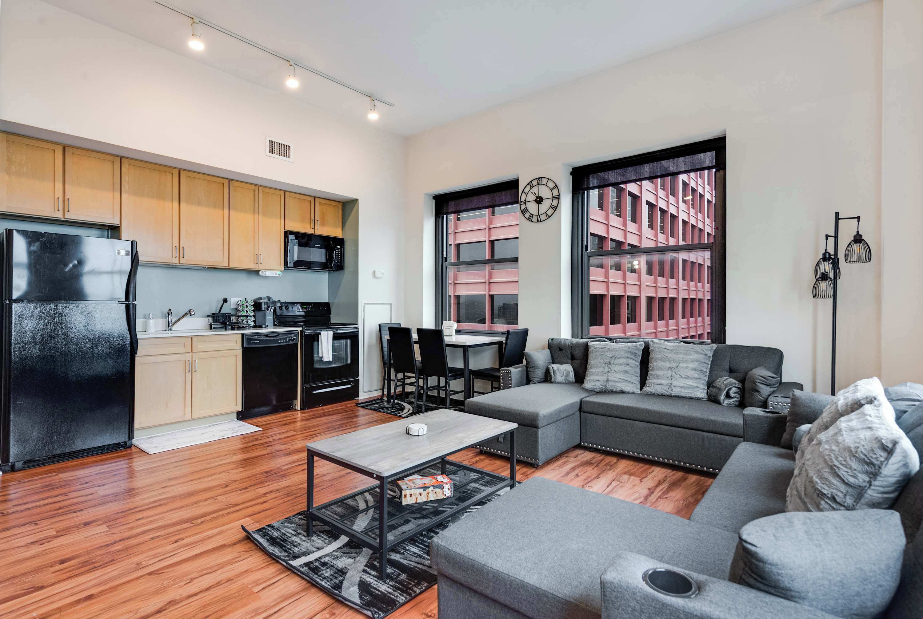 Un appartement moderne comprenant une cuisine, un coin repas et un salon. Il est doté de meubles de cuisine en bois, d’un réfrigérateur noir, d’une table à manger avec des chaises, d’un canapé gris et d’une grande fenêtre offrant une vue sur un immeuble.
