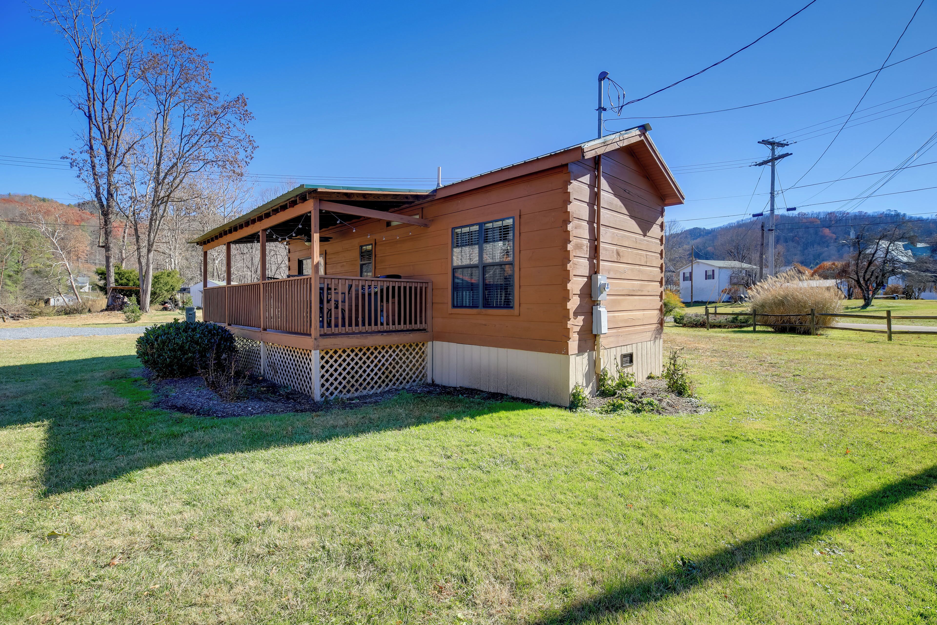 A single-story wooden cabin with a porch and a small deck, surrounded by a grassy yard and trees.