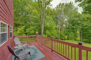 A red wooden deck with two Adirondack chairs and a table, surrounded by lush green trees.
