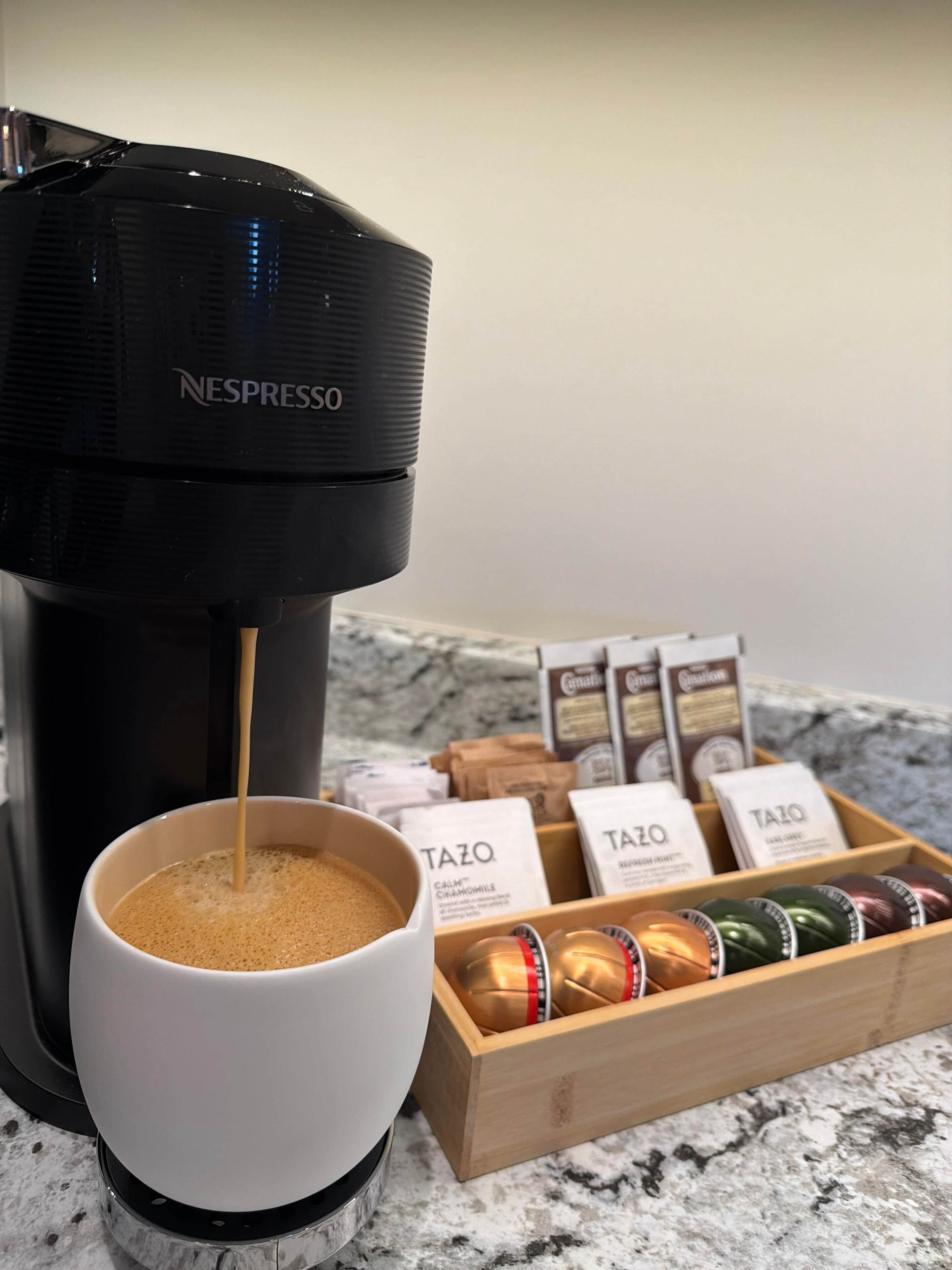 A Nespresso coffee machine dispensing coffee into a cup, with a box of Tazo coffee pods in the background.
