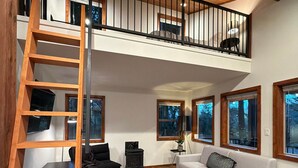 A spacious living area with a loft bedroom, wooden flooring, a white sofa, a black armchair, and a wooden ladder. - The Grove Guest House (Campbell River)