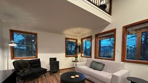 A modern living room with a high ceiling, wooden flooring, a black sofa, a white sofa, a black coffee table, and large windows with a view of trees. - The Grove Guest House (Campbell River)