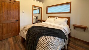 A bedroom with a wooden door, a bed with white bedding, a dark throw blanket, a woven basket, and a wooden shelf. - The Grove Guest House (Campbell River)