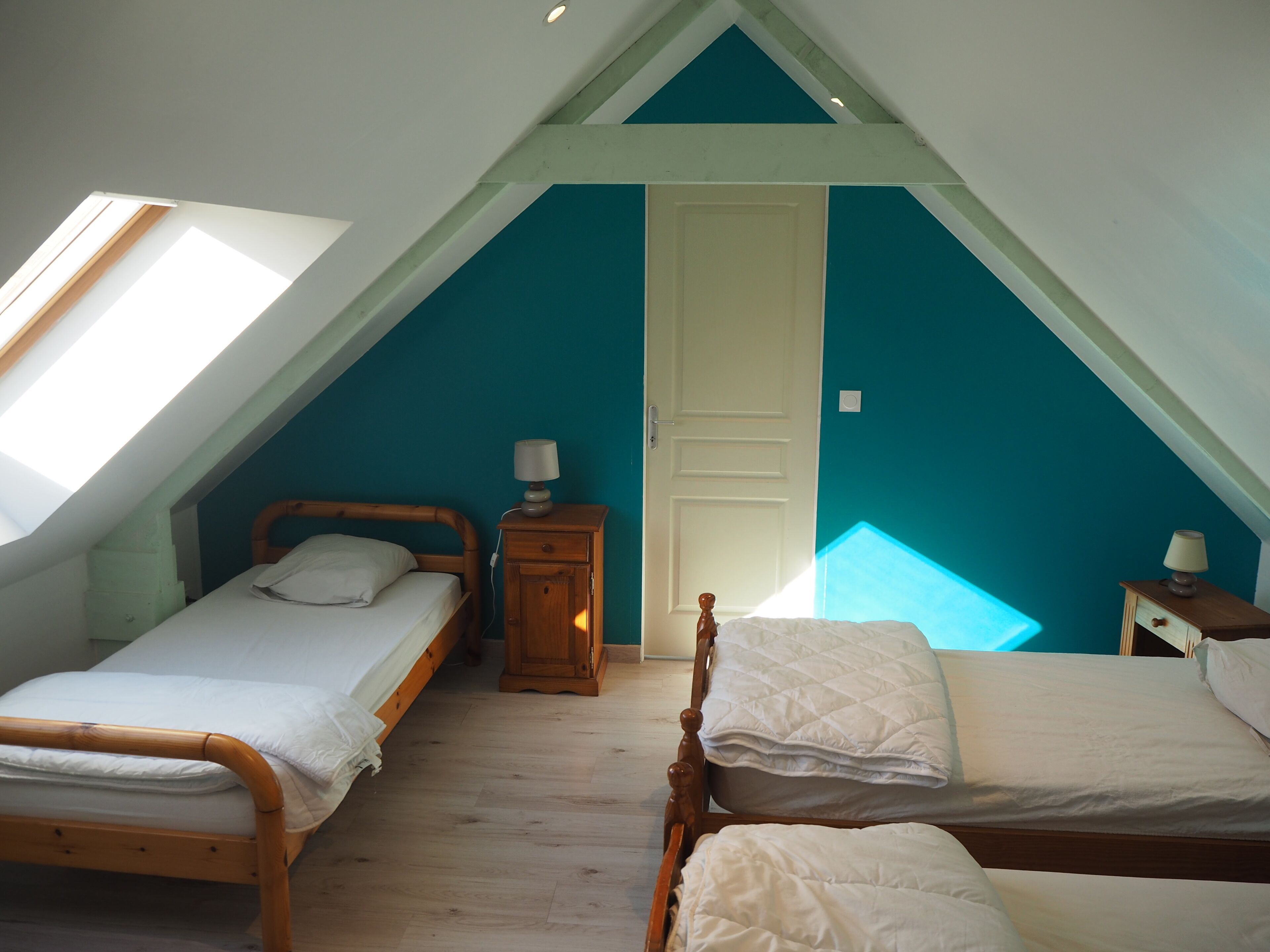 A room with two single beds, a wooden nightstand with a lamp, a door, and a skylight.