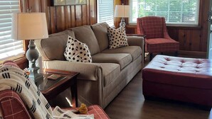 Een gezellige woonkamer met een houten lambrisering, een bank met opvallende kussens, een rode fauteuil en een rode poef.