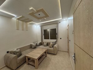 Un salon moderne avec un canapé d’angle, une table basse en bois, un éclairage encastré au plafond et une grande fenêtre agrémentée de rideaux.