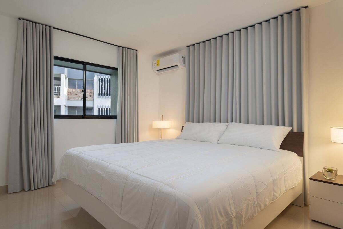 A hotel room with a large bed, a window with curtains, an air conditioning unit, a bedside table with a lamp, and a view of buildings outside.