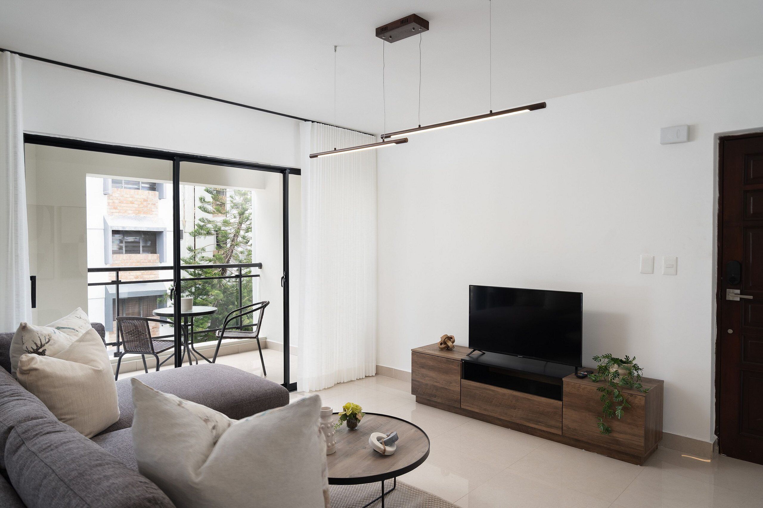 Una sala de estar moderna con un sofá, una mesa de centro, un televisor sobre un mueble de madera, y un balcón con una silla y una mesa.