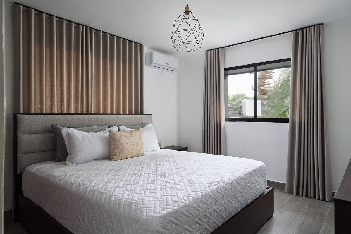 A modern bedroom with a large bed, a geometric pendant light, and a window with curtains.