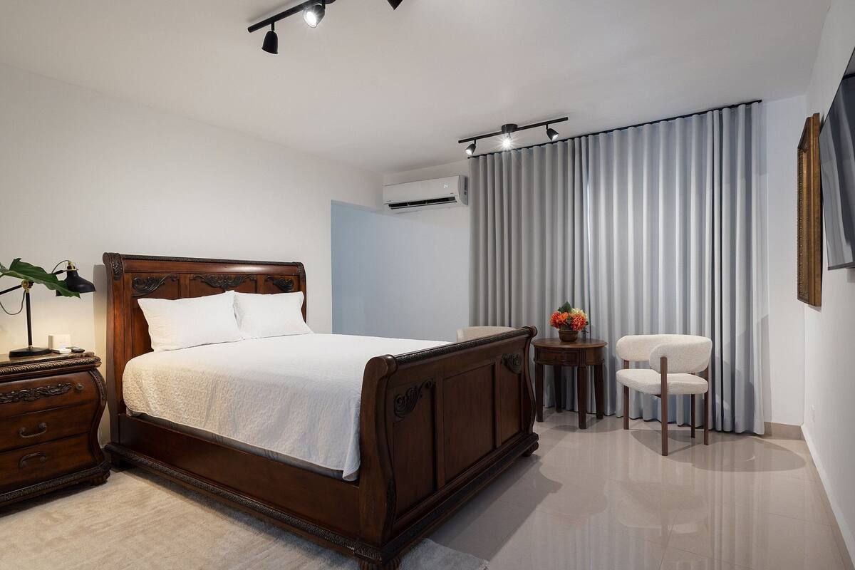 A neatly arranged bedroom with a wooden bed, a bedside table, a chair, and a small table with a vase.