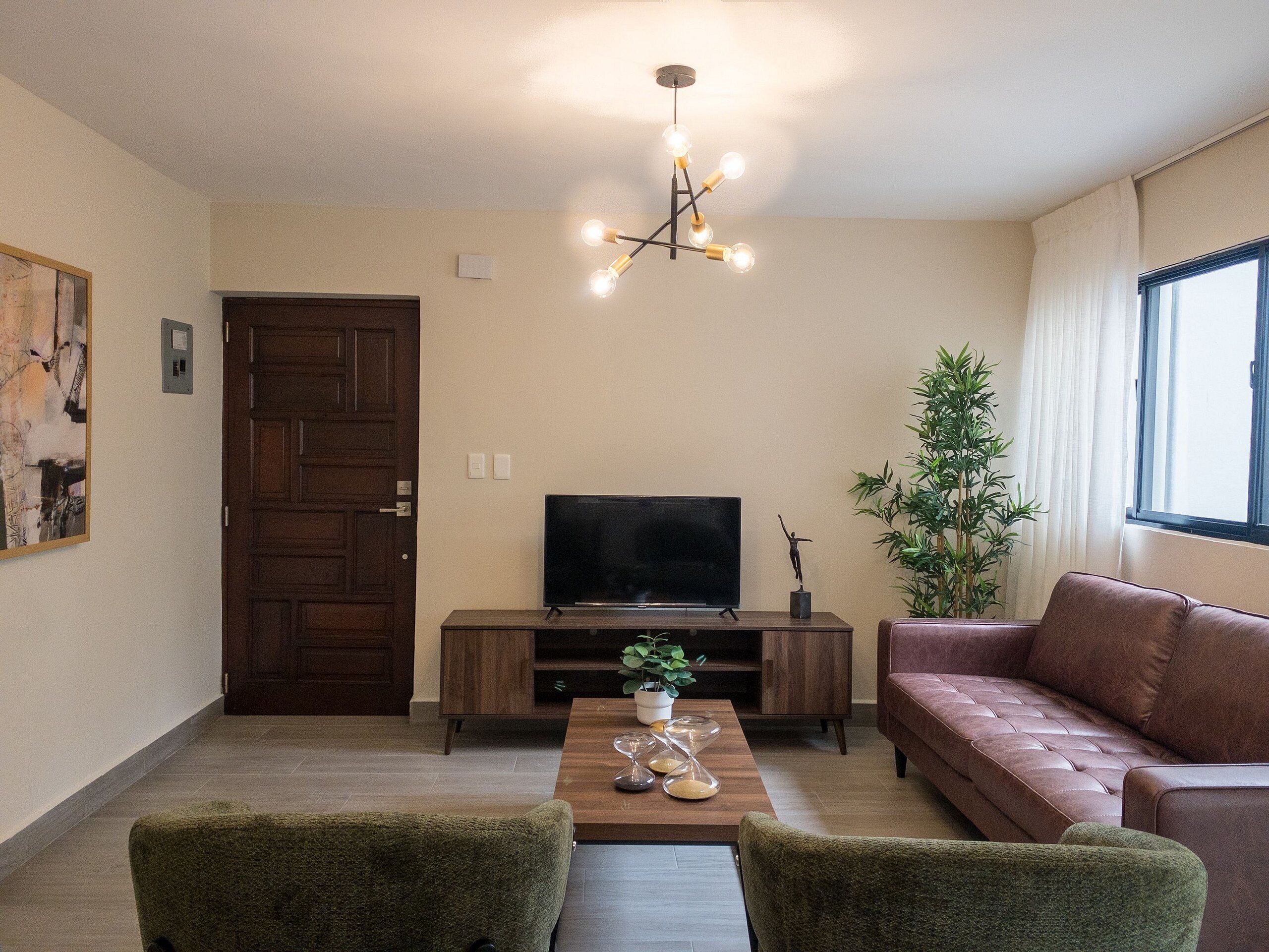 Ein Wohnzimmer mit Fernseher, Sofa, zwei Sesseln, einem Couchtisch mit Sanduhren, einer Pflanze und einer Tür.