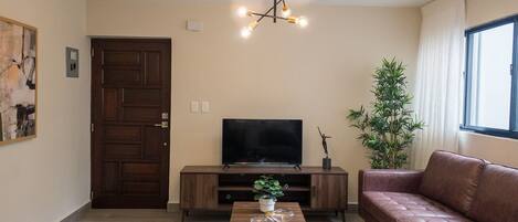 Ein Wohnzimmer mit Fernseher, Sofa, zwei Sesseln, einem Couchtisch mit Sanduhren, einer Pflanze und einer Tür.