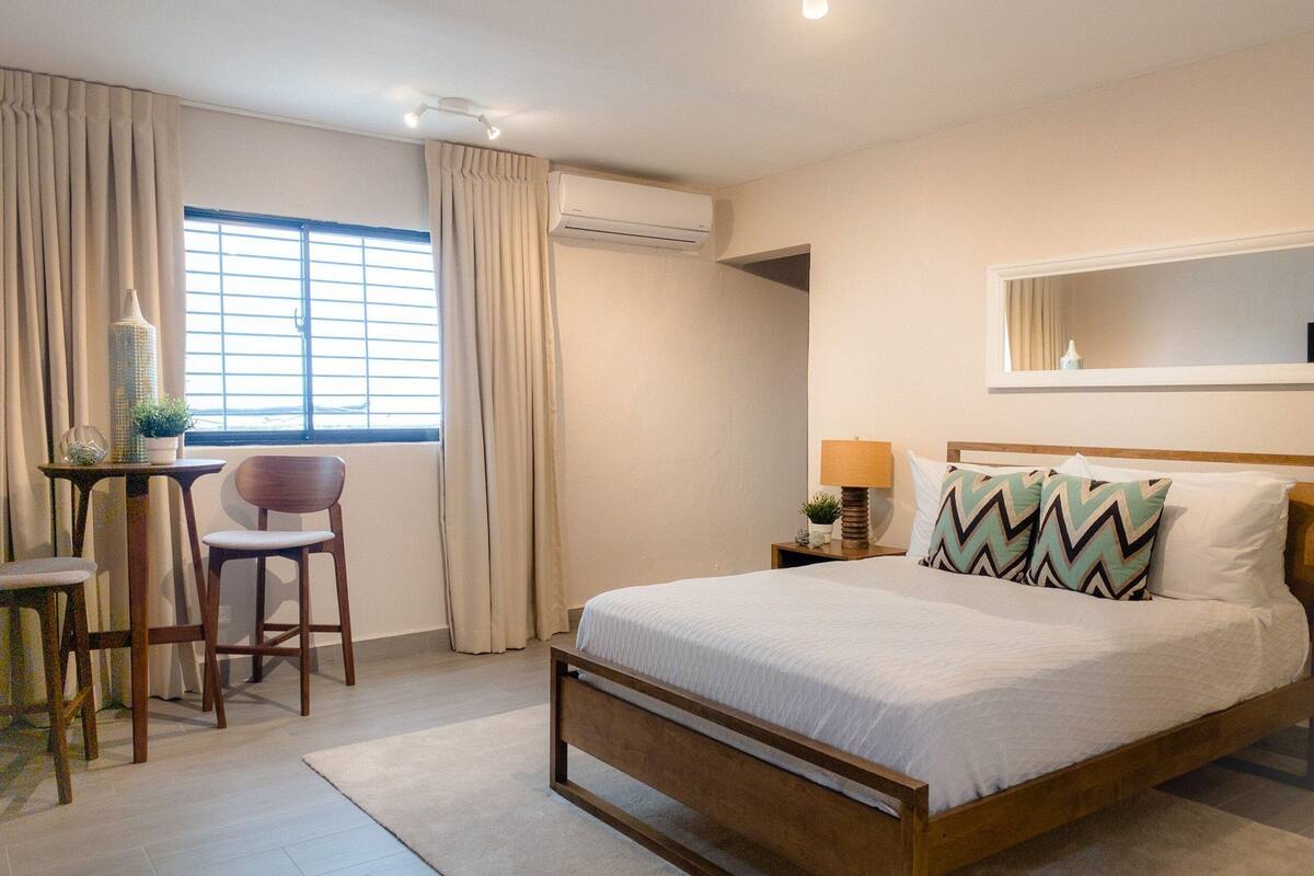 A bedroom with a wooden bed, a desk with a chair, a window with curtains, an air conditioning unit, and a bedside table with a lamp and decorative items.