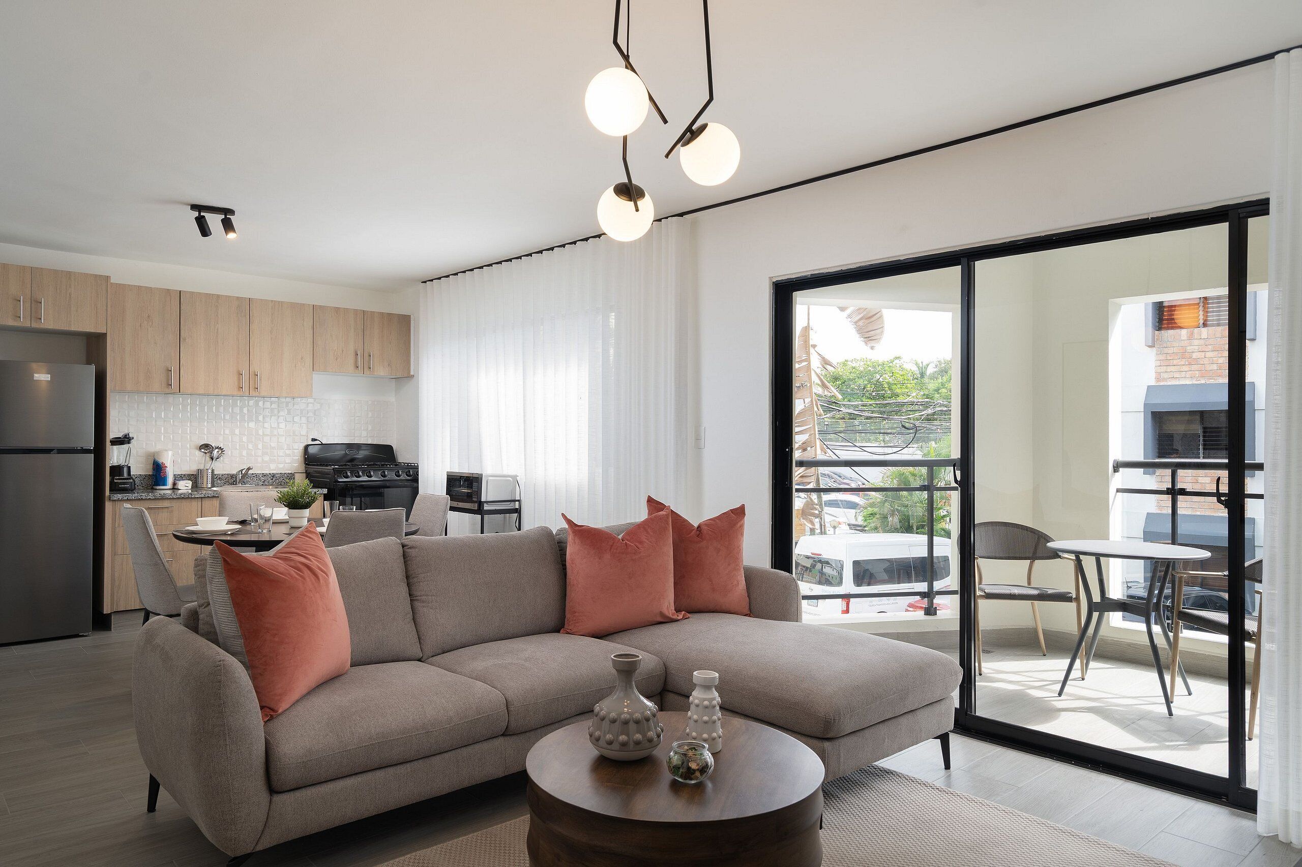 Un salon moderne avec un canapé gris, deux coussins roses et une table basse agrémentée de vases décoratifs. On aperçoit à l’arrière-plan une cuisine avec des meubles en bois et un coin repas composé d’une table et de chaises.