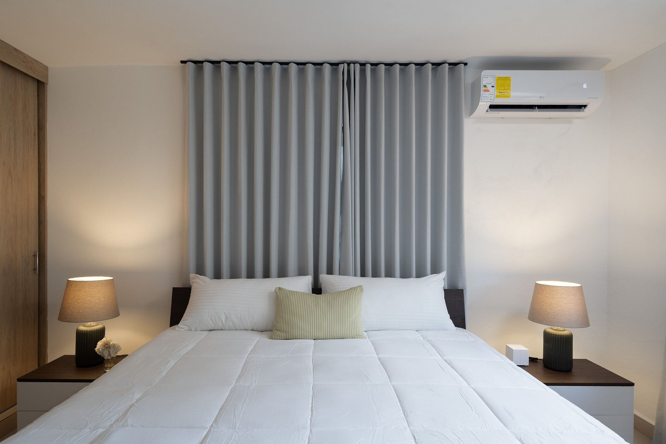 A hotel room with a bed, two bedside tables with lamps, a wall-mounted air conditioner, and grey curtains.