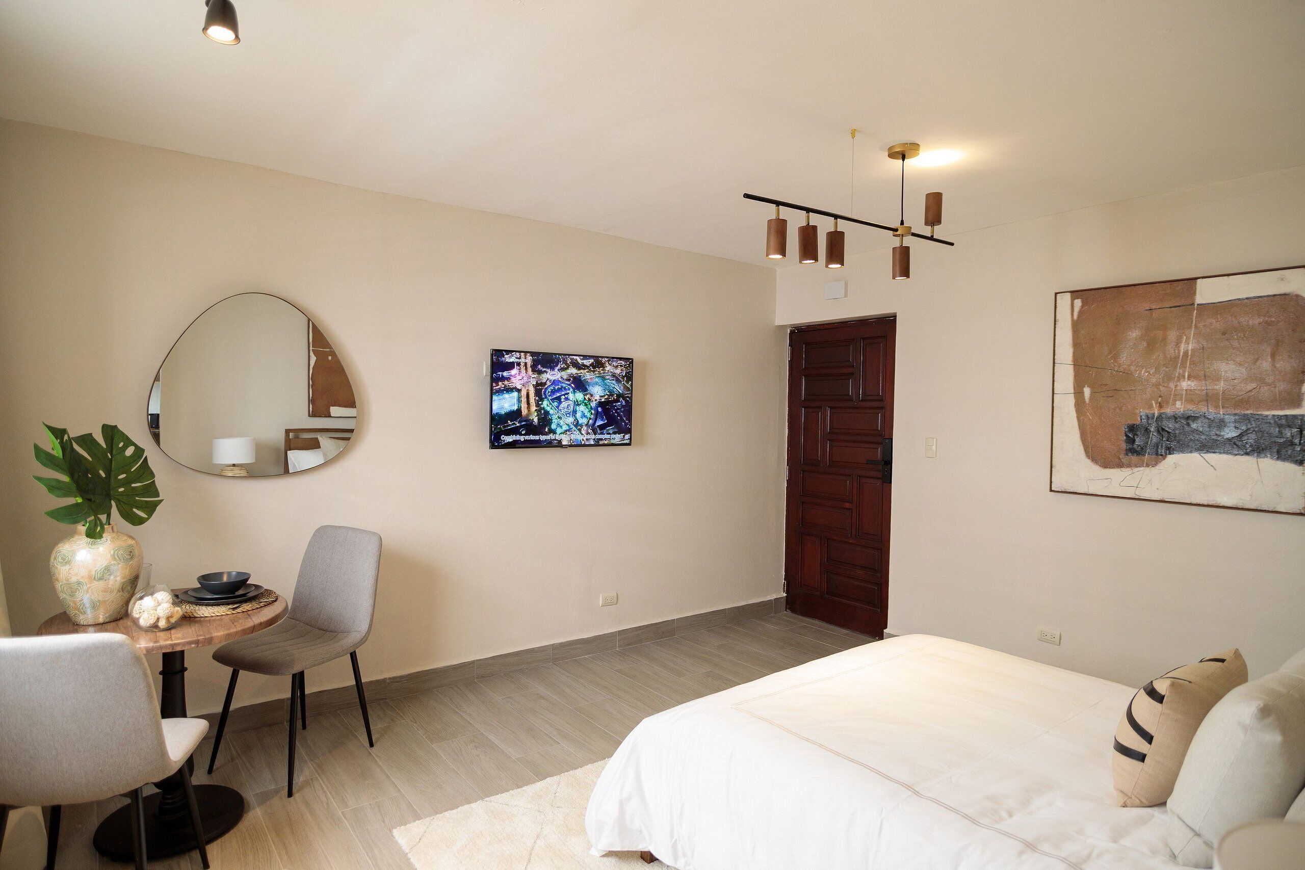 Een slaapkamer met een bed, een bureau met stoel, een ronde spiegel, een televisie en een houten deur.