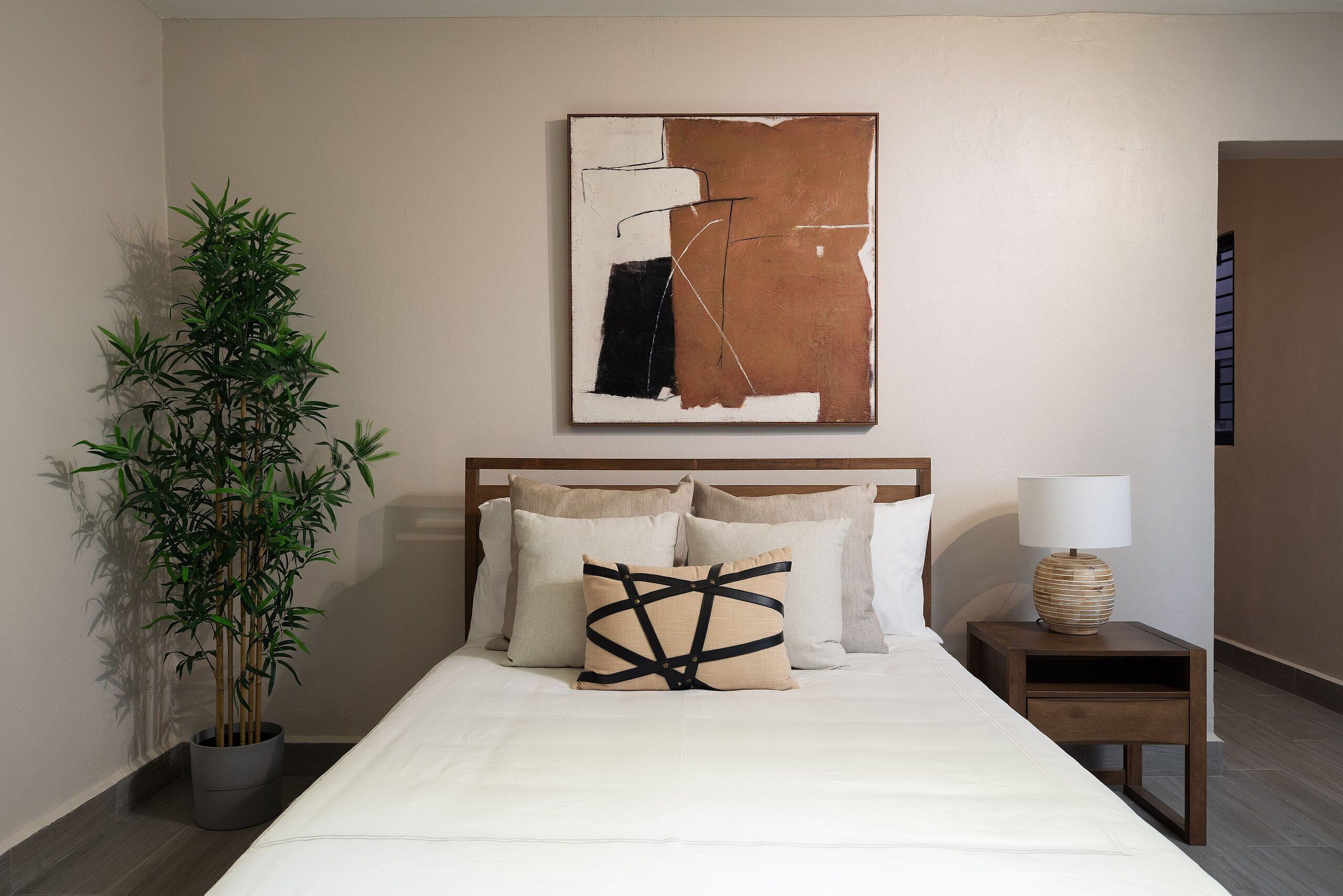 Un lit bien fait, avec une couette blanche et plusieurs oreillers, une table de chevet avec une lampe, et un grand tableau abstrait accroché au mur.