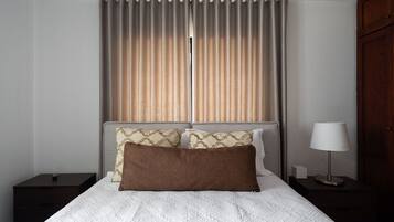 Una cama bien tendida con colcha blanca, dos almohadas, una lámpara de noche y una mesita de noche.