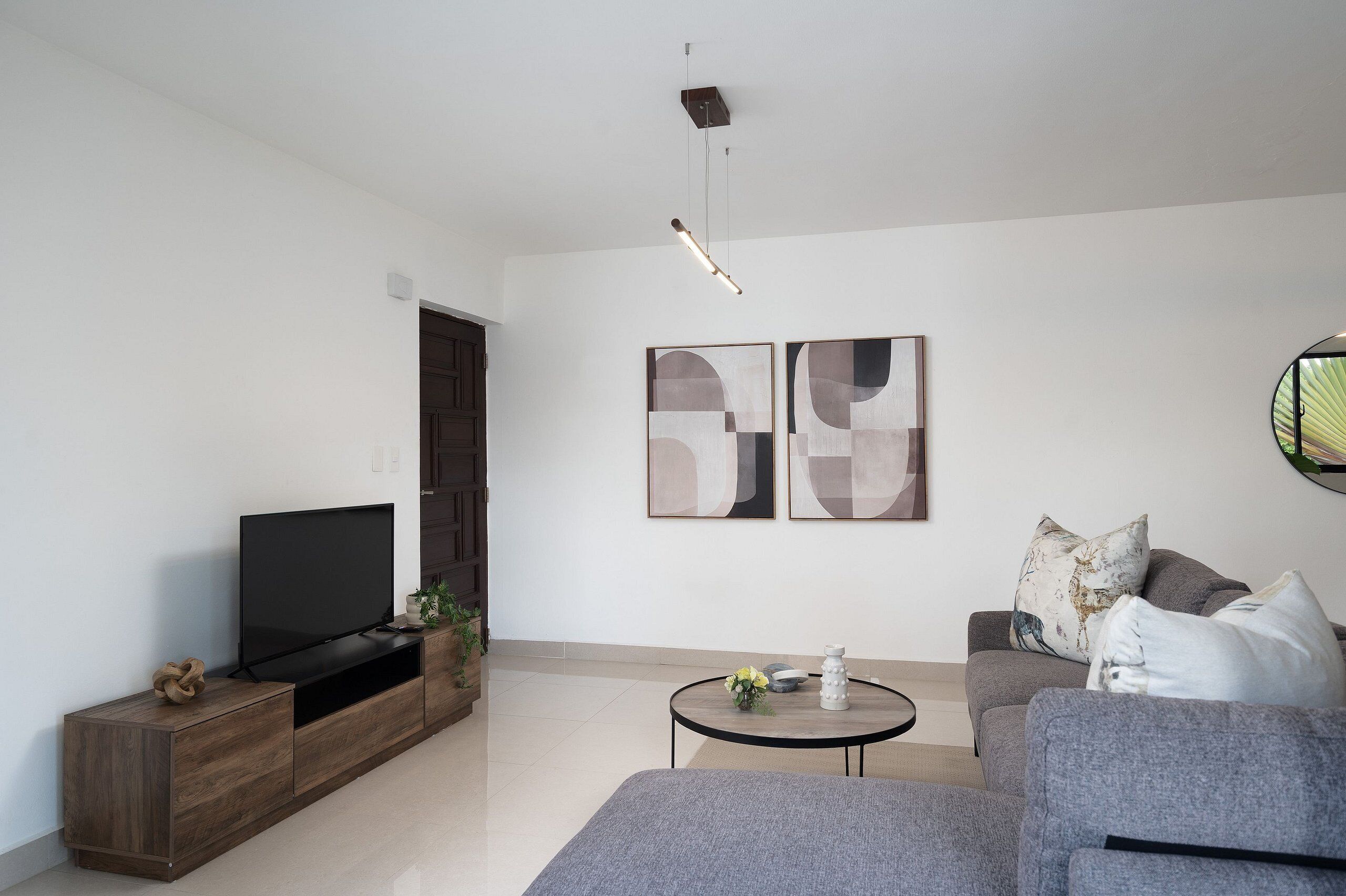 A modern living room with a flat-screen TV on a wooden TV stand, a grey sofa, a round coffee table with a vase, and abstract wall art.