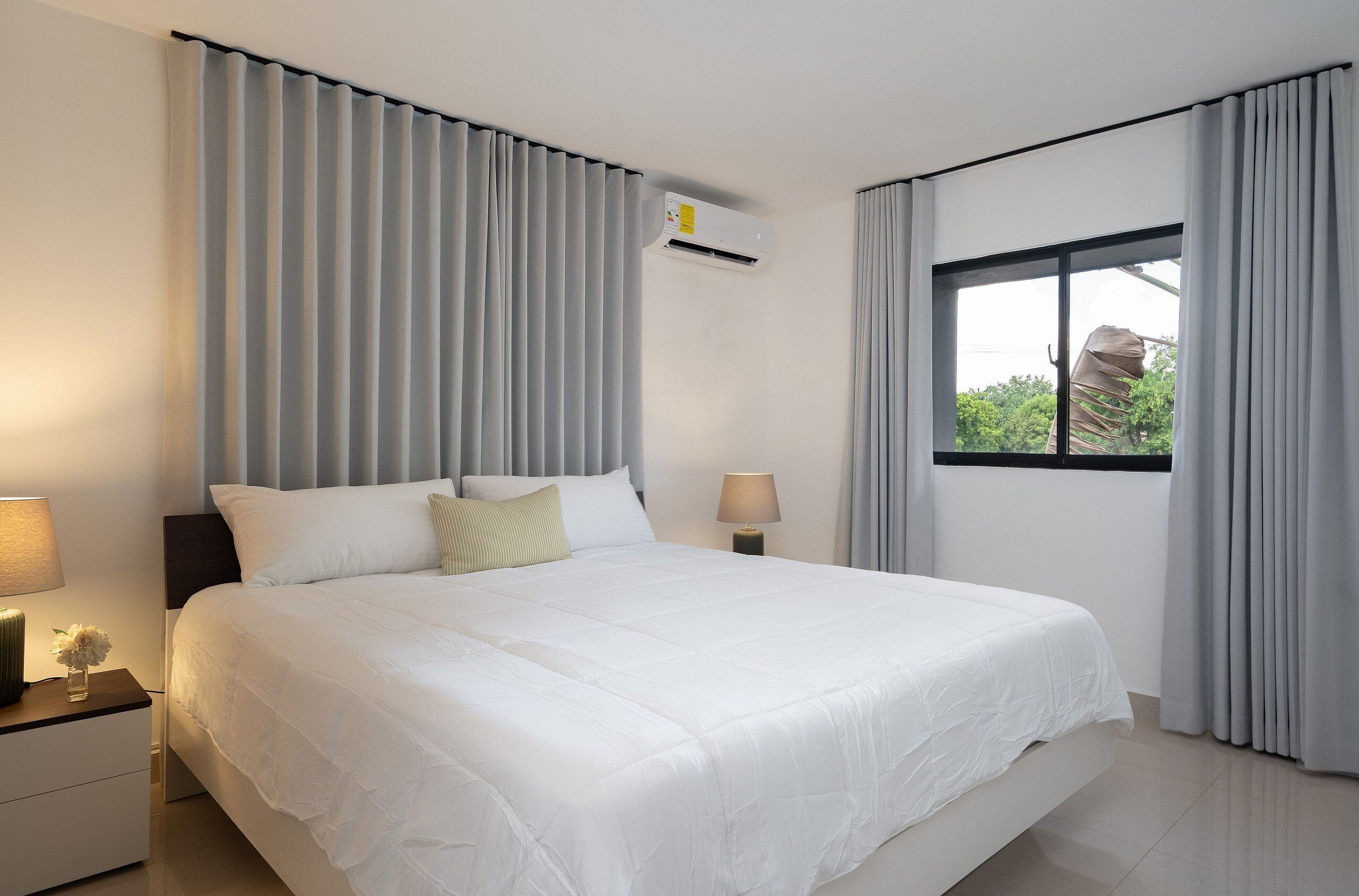 A modern hotel room with a large bed, a nightstand with a lamp, a window with curtains, and an air conditioning unit.