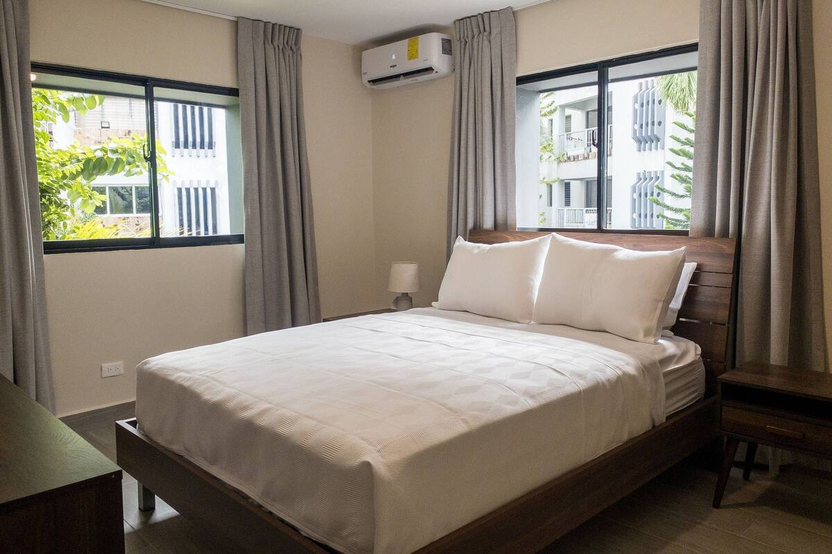 A neatly made bed with a wooden headboard, a bedside table, and a window with curtains.