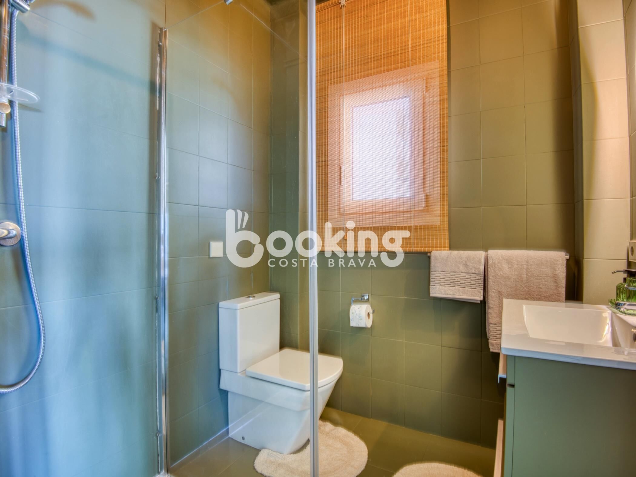 A bathroom with a glass shower enclosure, a toilet, a sink, and a window with blinds.