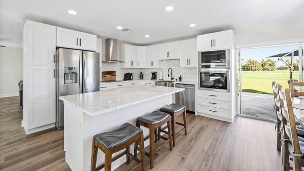 A modern kitchen with white cabinets, stainless steel appliances, a large island with bar stools, and a view of a green landscape through the glass door. - Golf & Beach Retreat Firepit BBQ Near Boca Grande (Rotonda West)