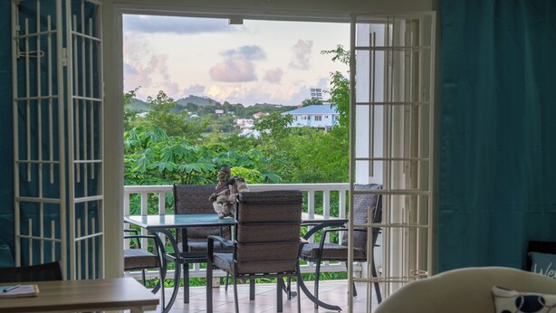 A room with a view of lush greenery and a distant house. There is a table with chairs and a sofa. - Sundrift Apartments (Gros Islet)